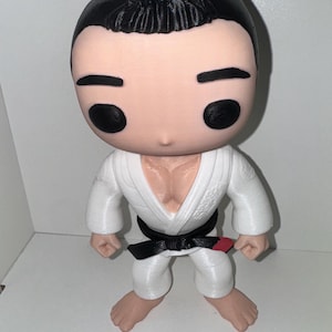 May include: A figurine of a martial artist in a white gi with a black belt and a red stripe. The figure has black hair and black eyes. The figurine is standing with its feet apart.