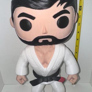 May include: A large, stylized figure of a martial artist in a white gi with a black belt and red accents. The figure has a black beard and hair, and is approximately 14 inches tall.