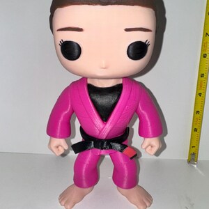 May include: A pink martial arts figure with a black belt and a black top. The figure has a brown hairstyle and large black eyes. The figure is standing upright, with its arms at its sides. A yellow measuring tape is in the background.