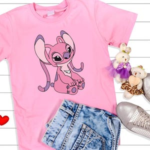May include: A pink t-shirt featuring a cartoon character in shades of pink and purple. The shirt is paired with denim shorts, silver sneakers, and two small stuffed animals. Two red heart-shaped decorations are also present.