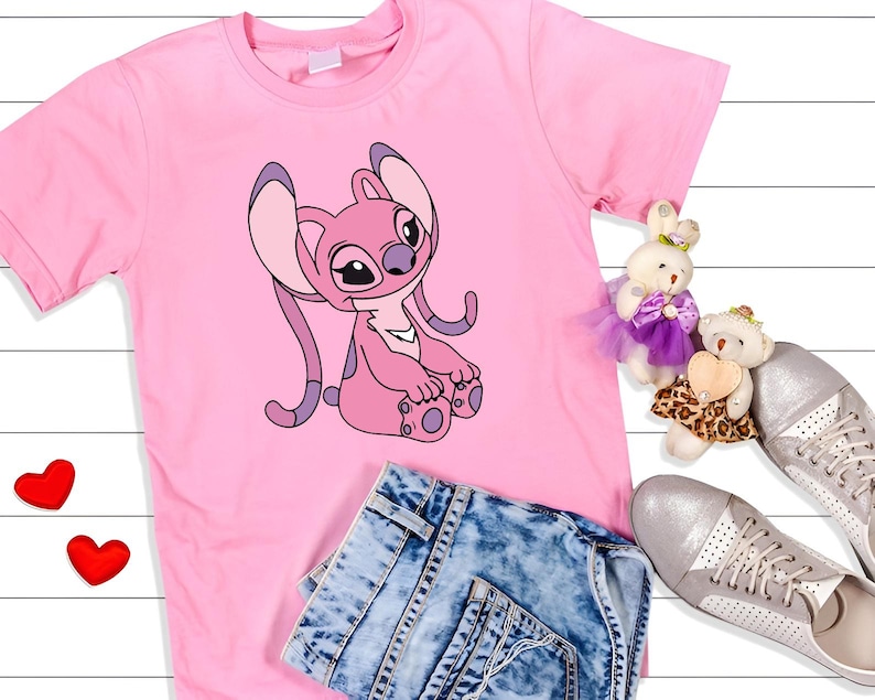 May include: A pink t-shirt featuring a cartoon character in shades of pink and purple. The shirt is paired with denim shorts, silver sneakers, and two small stuffed animals. Two red heart-shaped decorations are also present.