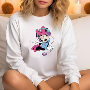 Może przedstawiać: Biała bluza z długim rękawem z kresk&oacute;wkowym wzorem Minnie Mouse. Myszka Minnie ma na sobie słuchawki i niebieski str&oacute;j. Grafika zawiera r&oacute;żowe, fioletowe i czarne akcenty. Widoczny jest r&oacute;wnież mały odtwarzacz muzyki.