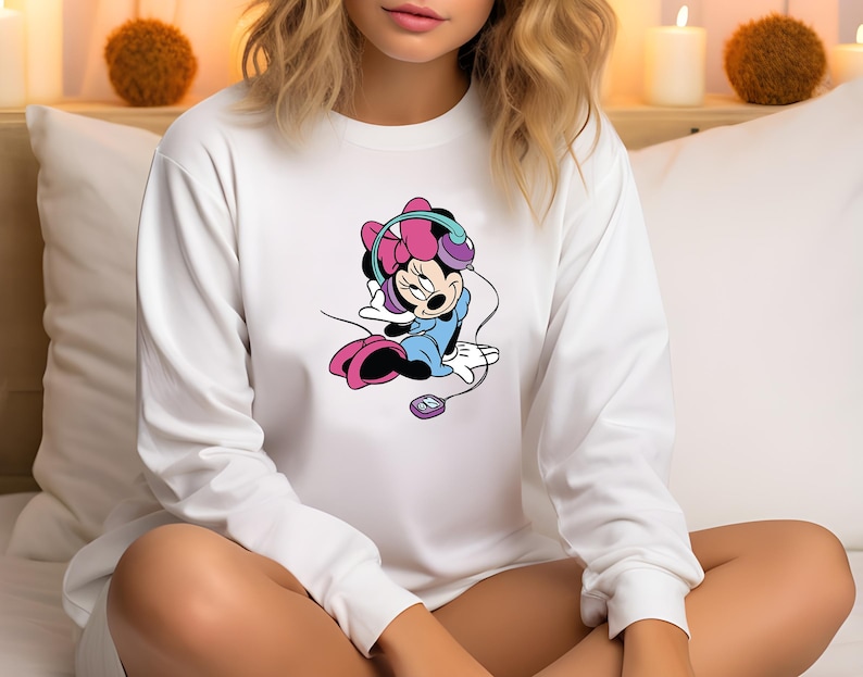 Może przedstawiać: Biała bluza z długim rękawem z kresk&oacute;wkowym wzorem Minnie Mouse. Myszka Minnie ma na sobie słuchawki i niebieski str&oacute;j. Grafika zawiera r&oacute;żowe, fioletowe i czarne akcenty. Widoczny jest r&oacute;wnież mały odtwarzacz muzyki.