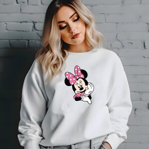 Może przedstawiać: Biała bluza z nadrukiem Myszki Minnie. Grafika przedstawia Myszkę Minnie w r&oacute;żowej sukience i kokardzie w białe grochy. Swobodny, wygodny styl ubioru.