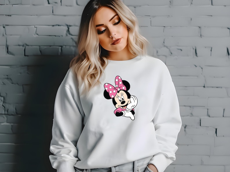Może przedstawiać: Biała bluza z nadrukiem Myszki Minnie. Grafika przedstawia Myszkę Minnie w r&oacute;żowej sukience i kokardzie w białe grochy. Swobodny, wygodny styl ubioru.