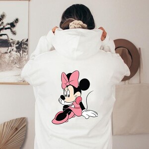 Może przedstawiać: Biała bluza z kapturem z grafiką Myszki Minnie na plecach. Grafika przedstawia Myszkę Minnie w r&oacute;żowej sukience i kokardzie. Bluza ma kaptur ze sznurkiem i kieszeń typu kangur.