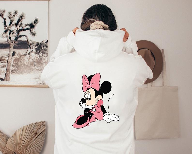 Może przedstawiać: Biała bluza z kapturem z grafiką Myszki Minnie na plecach. Grafika przedstawia Myszkę Minnie w r&oacute;żowej sukience i kokardzie. Bluza ma kaptur ze sznurkiem i kieszeń typu kangur.