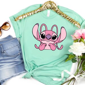 May include: A mint green t-shirt featuring a pink cartoon character with large ears and a purple nose. The shirt is displayed on a gold sequined hanger, next to a pair of blue denim shorts, sunglasses, and a bouquet of pink and white flowers.