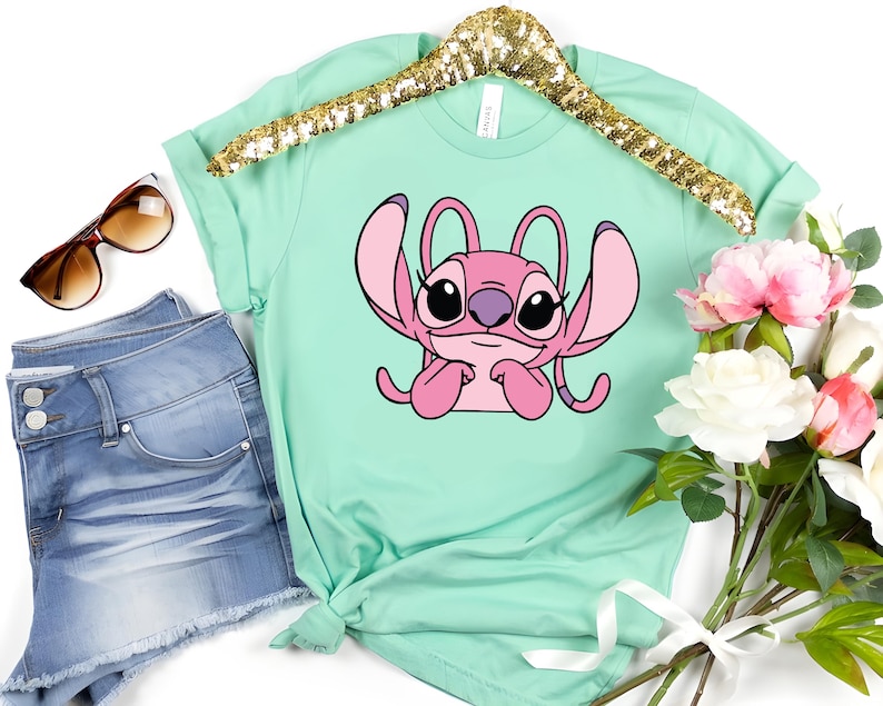 May include: A mint green t-shirt featuring a pink cartoon character with large ears and a purple nose. The shirt is displayed on a gold sequined hanger, next to a pair of blue denim shorts, sunglasses, and a bouquet of pink and white flowers.