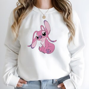 May include: White sweatshirt featuring a pink cartoon character with large ears and a seated pose. The character has pink and purple accents. The sweatshirt has long sleeves and a crew neck.