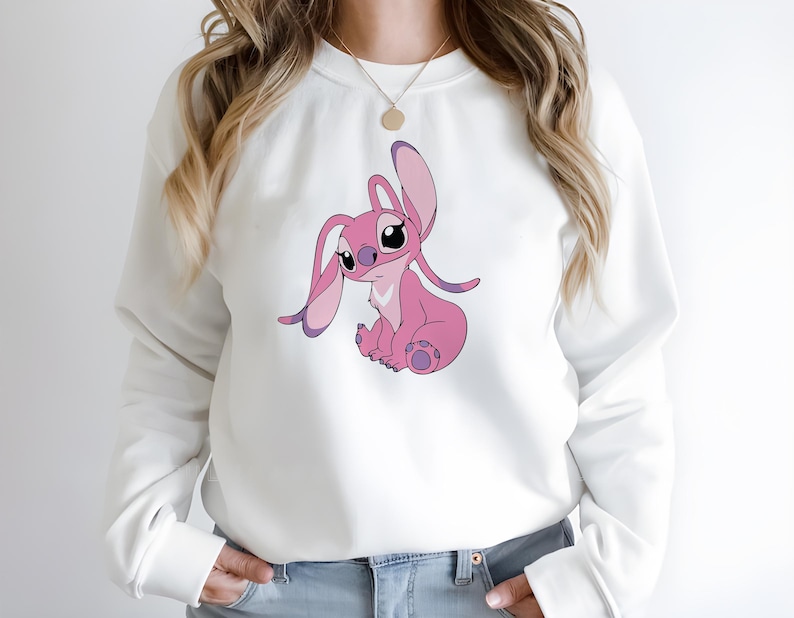 May include: White sweatshirt featuring a pink cartoon character with large ears and a seated pose. The character has pink and purple accents. The sweatshirt has long sleeves and a crew neck.