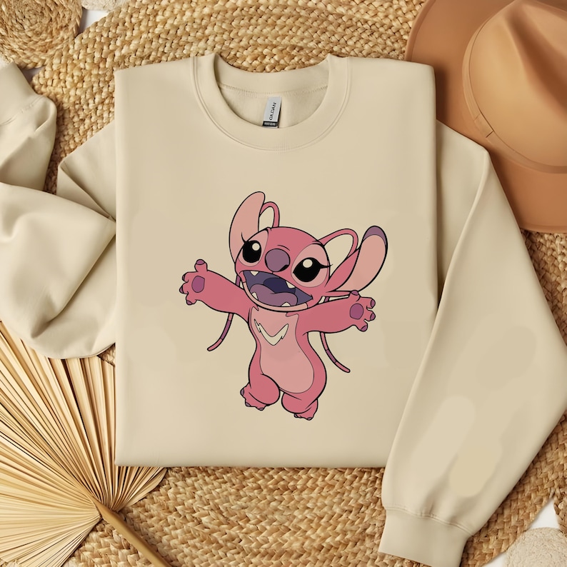 May include: A cream-colored sweatshirt featuring a pink cartoon character with large ears and a welcoming pose. The character has a light pink body, darker pink accents, and a wide, open mouth. The sweatshirt has a crew neck and long sleeves.