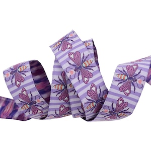 May include: A lavender ribbon with a repeating pattern of purple bees and white stripes. The bees have pink hearts and orange and white striped abdomens. The ribbon is curled and folded, showing a glimpse of a purple and pink patterned fabric.
