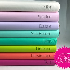 May include: A stack of eight fabric rolls in various colors, including white, pink, lavender, mint green, teal, lime green, coral, and magenta. The fabric rolls are labeled with the names "Mist", "Sparkle", "Dazzle", "Sea Breeze", "Julep", "Limeade", "Persimmon", and "Tula". The Tula Pink logo is visible in the bottom right corner.