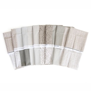 May include: A collection of eight folded fabric pieces arranged in a fan shape, featuring various patterns in shades of beige, taupe, and white. Each piece is secured with a white paper band labeled Mod Texture, showcasing geometric designs.