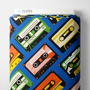 May include: A bolt of fabric featuring a repeating pattern of colorful cassette tapes on a bright blue background. The tapes are depicted in various shades of green, orange, yellow, and black, with some labeled as 60 or 90. The top edge shows a white label with the text CLOUD9.