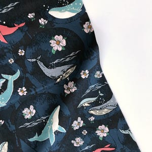 May include: Dark blue fabric with a repeating pattern of whales in shades of blue and pink, along with white and pink flowers. The whales are swimming in a sea of dark blue circles.