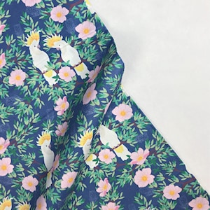 May include: A blue fabric with a repeating pattern of white cockatoos perched on branches with pink flowers. The cockatoos have yellow crests and are surrounded by green leaves.