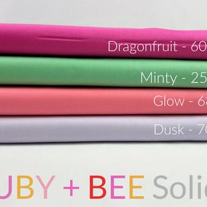 May include: Four rolls of solid-coloured fabric in shades of pink, green, coral, and lavender. Each roll is labelled with a colour name and number. The text "RUBY + BEE Solids" is displayed at the bottom.
