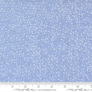 May include: A light blue fabric featuring a white speckled pattern of small dots and a subtle grid-like texture. A white ruler is visible at the bottom edge, showing measurements in inches, with the text moda and www.modafabrics.com printed.