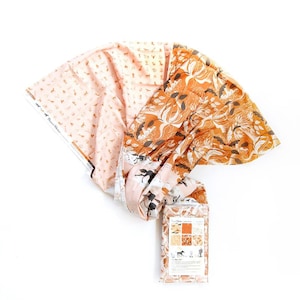 May include: A bundle of fabric featuring two distinct patterns. One section is light pink with small brown desert-themed icons. The other section is a vibrant orange with a large-scale white botanical print. A rectangular product label is attached.