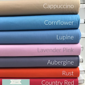Peut inclure: Un ensemble de sept échantillons de tissu de différentes couleurs, dont cappuccino, bleuet, lupin, rose lavande, aubergine, rouille et rouge campagnard. Les échantillons sont étiquetés avec leurs couleurs respectives. Le logo Tilda Solids est visible en bas de l'image.