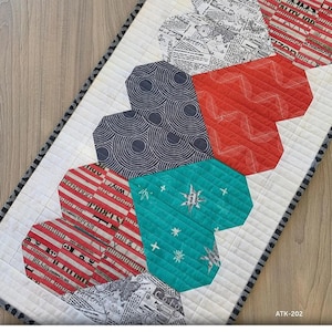May include: A quilt pattern for a table runner featuring a row of hearts in various colours and patterns. The text "Let's Play HEARTS!" is at the top of the runner. The pattern is called "ATK-202".