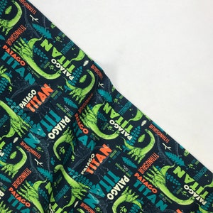 May include: A dark blue fabric with a repeating pattern of green dinosaurs and the text "Patago Titan" in orange and white. The dinosaurs are outlined in green and have a light green body. The fabric has a subtle texture.