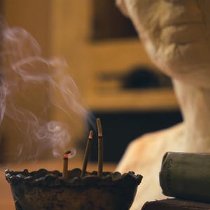 May include: A bronze incense burner with three burning incense sticks, emitting wisps of smoke. The burner has a scalloped rim and three small feet. A bust and books are in the background.