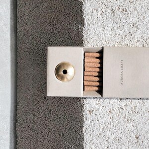 May include: A light grey box containing brown incense sticks and a brass incense holder. The box is labelled "AURORA CRAFT". The background is a textured grey surface. The image is well-lit, highlighting the product.