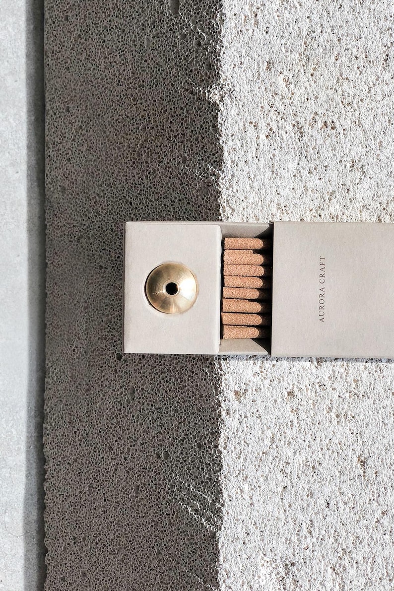 May include: A light grey box containing brown incense sticks and a brass incense holder. The box is labelled "AURORA CRAFT". The background is a textured grey surface. The image is well-lit, highlighting the product.