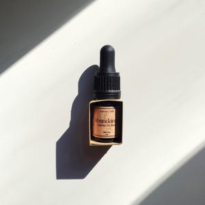 Könnte beinhalten: Kleine Flasche mit ätherischem Öl-Gemisch mit schwarzer Pipette und Etikett mit der Aufschrift "Abundance Essential Oil Blend". Die Flasche ist braun mit einer goldfarbenen Box. Der Text "Aurora Craft" steht ebenfalls auf dem Etikett. Die Flasche enthält 5 ml Öl.