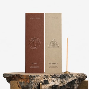May include: Two rectangular cardboard boxes of incense sticks, one dark brown labelled Gaia and one light beige labelled Shaman, both featuring Aurora Craft branding and embossed symbols. A single thin wooden incense stick stands upright nearby.