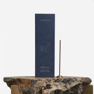 Pu&ograve; includere: Una scatola blu navy di bastoncini di incenso con il testo "AURORA CRAFT" e "RITUAL 100% NATURAL INCENSE STICKS". Un singolo bastoncino di incenso &egrave; in piedi accanto alla scatola. La scatola e l'incenso sono su una superficie di pietra.
