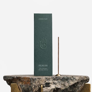 Può includere: Una scatola verde scuro di bastoncini di incenso naturali "HEALER" 100% con un simbolo a mano in rilievo sul davanti. Un singolo bastoncino di incenso è in posizione verticale in un piccolo supporto in ottone. La scatola e l'incenso sono esposti su una superficie di pietra.