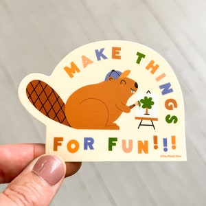 Könnte beinhalten: Ein Aufkleber mit einem Cartoon-Biber, der einen blauen Hut trägt und einen grünen Baum auf eine Leinwand malt. Der Aufkleber lautet "MAKE THINGS FOR FUN!!!" in Regenbogenfarben.