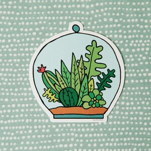 May include: A sticker of a glass terrarium with a variety of green plants, including a cactus, succulents, and ferns. The terrarium is on a light blue background with a white border.