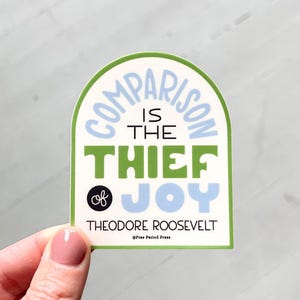 May include: A sticker with a green border and a light blue and green design. The text reads "COMPARISON IS THE THIEF OF JOY" with the name "THEODORE ROOSEVELT" below. The sticker is held by a person.