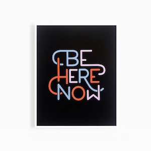Op de afbeelding: Een zwart-witte print met de tekst "BE HERE NOW" in kleurrijke, overlappende letters. De letters zijn in tinten blauw, roze en oranje.