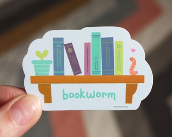 Peut inclure: Un autocollant blanc avec une illustration de dessin animé d'une étagère avec des livres colorés, une plante en pot et un ver. Le texte "bookworm" est écrit sous l'étagère.