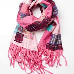 May include: A patchwork scarf featuring various fabric squares in shades of pink, purple, and teal, including plaid and floral patterns. The scarf is constructed with visible decorative stitching and finished with long, frayed pink fabric fringe ends.