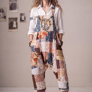 May include: Patchwork jumpsuit with a deer graphic on the bib. The jumpsuit features a variety of floral and geometric patterns in shades of blue, brown, and cream. The model is wearing a white shirt and brown boots.