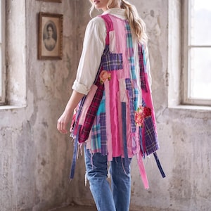 May include: A sleeveless vest featuring a patchwork design of pink, blue, and purple plaid fabric strips. The garment includes frayed edges, hanging fabric ribbons, and floral appliques. It is worn over a white long-sleeve shirt and blue jeans.