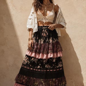 May include: A long tiered skirt featuring a black base with floral patterns, alternating with solid dusty pink fabric panels and dark lace trim. The skirt has a drawstring waist with tassels and is paired with a cream lace bralette and white jacket.