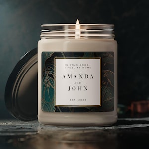 Puede incluir: Una vela blanca con una etiqueta negra y dorada que dice "En tus brazos, me siento en casa. Amanda y John Est. 2023". La etiqueta tiene un diseño de hojas tropicales.