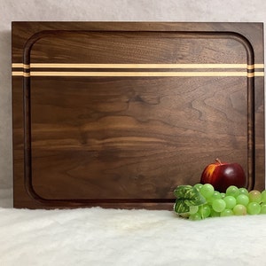May include: A large, rectangular walnut cutting board with a rounded edge and two thin, light brown wood strips inlaid across the top. The board is sitting on a white surface with a red apple and a bunch of green grapes in front of it.