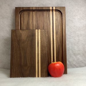 May include: Two wooden cutting boards with a walnut finish and maple stripes. The smaller board is in front of the larger board. A red apple is in front of the boards.