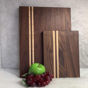 May include: Two wooden cutting boards, one rectangular and one square, are displayed. Both are dark brown with two light-colored wood stripes down the center. A green apple and a bunch of red grapes are in front of the boards.