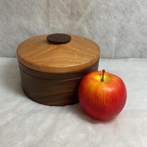 May include: A round wooden box with a lid, featuring a light brown top and a darker brown base. A small, dark brown knob sits atop the lid. A red and yellow apple is next to the box.