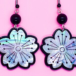 Peut inclure: Une paire de boucles d'oreilles florales avec un contour noir et un centre irisé. Chaque boucle d'oreille comporte une perle noire et un fil argenté. Les boucles d'oreilles sont posées sur un fond rose.